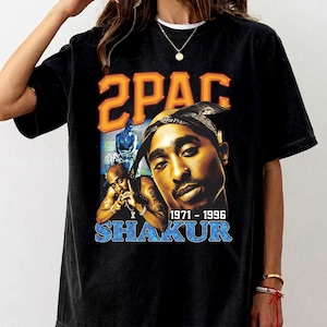 May include: A black short-sleeve t-shirt featuring a graphic print of Tupac Shakur. The design includes the text 2PAC in orange, SHAKUR in blue, and the years 1971-1996. The graphic displays a large portrait of Tupac and a smaller image of him.