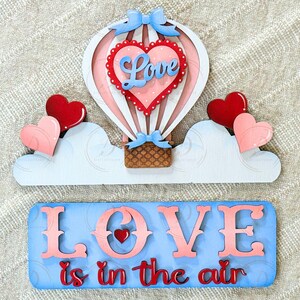 May include: A wooden Valentine's Day decoration featuring a hot air balloon with a heart that says "Love". Two red hearts and two pink hearts are on either side of the balloon. The phrase "Love is in the air" is below.