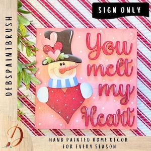 May include: A hand-painted wooden sign featuring a snowman with a top hat, scarf, and heart-shaped body. The sign reads "You melt my Heart" in red script. The background is pink with white polka dots, and the sign is framed with a red and white striped border.