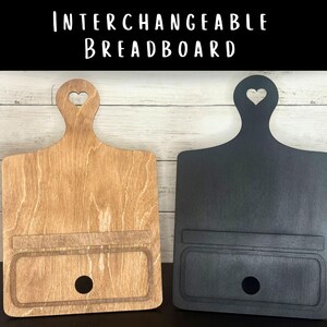 May include: Two wooden breadboards, one natural and one black, with a handle featuring a heart cutout. Each board has a rectangular inset and a circular hole. The text "INTERCHANGEABLE BREADBOARD" is at the top.