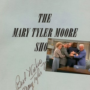 May include: A black and white photo of the cast of "The Mary Tyler Moore Show" hugging each other. The photo is on a blue script page with the title "The Mary Tyler Moore Show" and the text "The Last Show" and "PROD. #6027". The script page also has the signatures of Mary Tyler Moore and Ed Asner.
