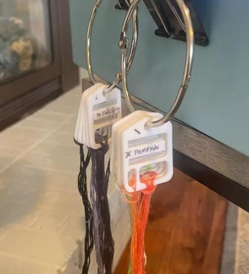 May include: Two white plastic embroidery floss organizers held on metal binder rings. Each organizer features handwritten labels reading "Dk Slate Grey" and "3K Pumpkin" with corresponding dark grey and orange embroidery threads attached to the slots.