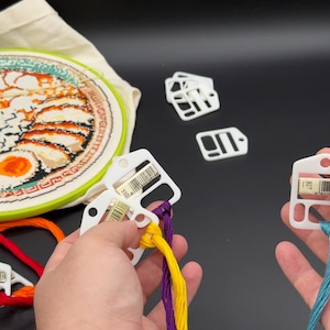 May include: A round embroidery hoop containing a fabric piece with a ramen bowl design, accompanied by several white plastic thread bobbins. Some bobbins are attached to colorful embroidery floss, while others are loose or on a metal ring.