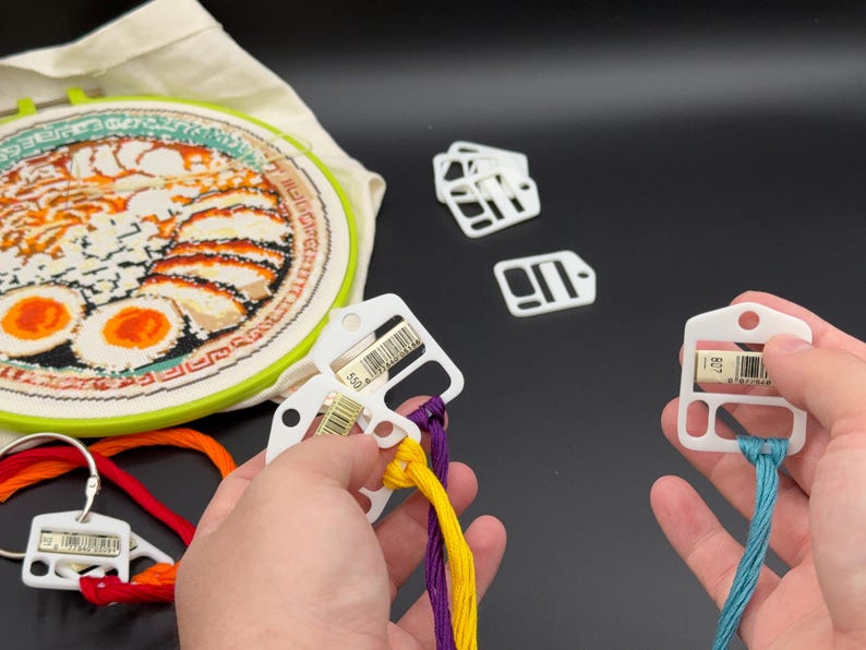 May include: A round embroidery hoop containing a fabric piece with a ramen bowl design, accompanied by several white plastic thread bobbins. Some bobbins are attached to colorful embroidery floss, while others are loose or on a metal ring.