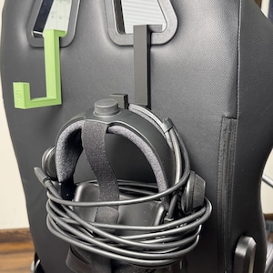 May include: Black VR headset with attached cables hanging on a black chair. A black and a green headset holder are attached to the chair. The headset has a black strap and gray padding.
