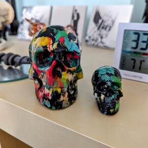 May include: Two decorative skull figurines. One skull is black with colorful paint splatters, the other is black with green and white floral designs.