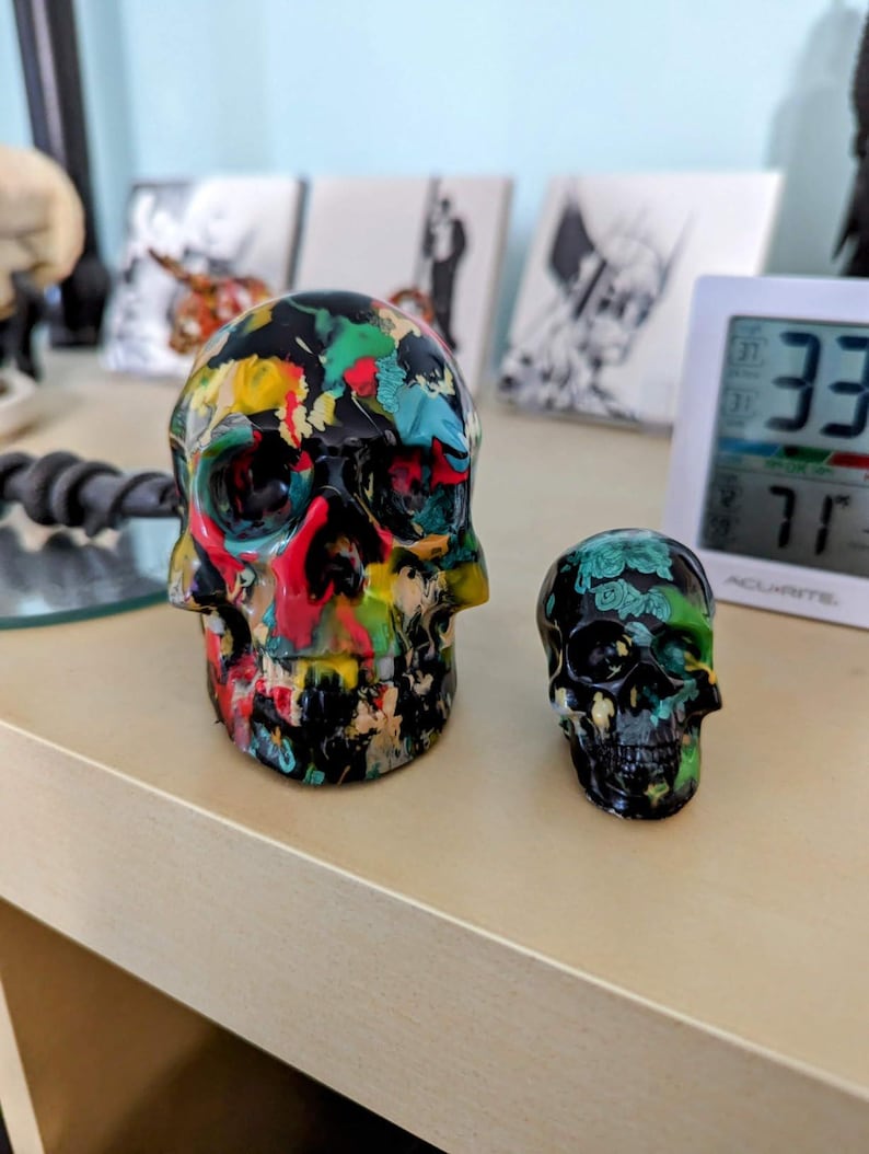 May include: Two decorative skull figurines. One skull is black with colorful paint splatters, the other is black with green and white floral designs.