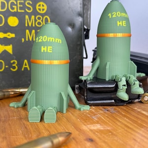 Puede incluir: Dos objetos verdes en forma de proyectiles de artillería, cada uno con el texto "120mm HE" y una banda naranja. Uno está sobre un objeto negro, el otro sobre una superficie de madera. Munición y una caja metálica verde en el fondo.