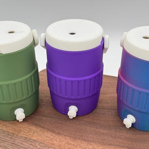 May include: Three colorful beverage coolers in green, purple, and blue with white lids and spouts. Each cooler has a ribbed design around the middle and a white handle. The coolers are displayed on a wooden surface.