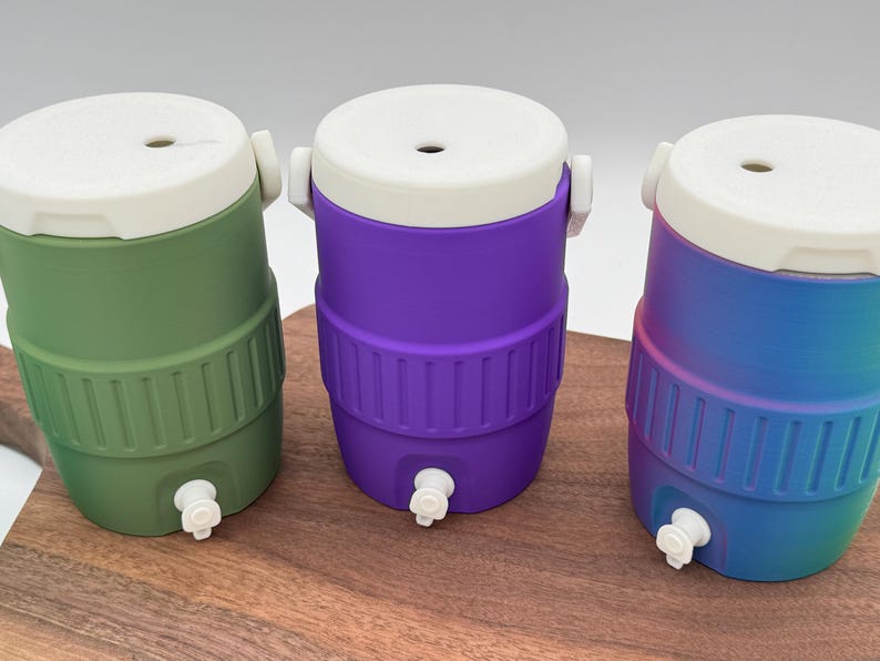 May include: Three colorful beverage coolers in green, purple, and blue with white lids and spouts. Each cooler has a ribbed design around the middle and a white handle. The coolers are displayed on a wooden surface.