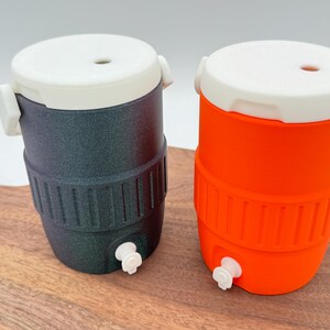 May include: Two small, insulated beverage coolers. One is dark gray with a white lid and accents, the other is bright orange with a white lid and accents. Both have a spigot at the bottom and are sitting on a wooden surface.