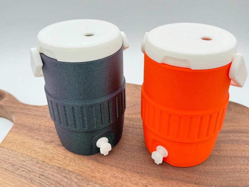 May include: Two small, insulated beverage coolers. One is dark gray with a white lid and accents, the other is bright orange with a white lid and accents. Both have a spigot at the bottom and are sitting on a wooden surface.