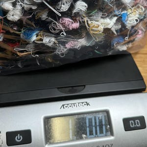 May include: A digital scale with a black base and a silver top. The scale has a digital display that reads "0.0". A clear plastic bag filled with colorful yarn is on top of the scale.
