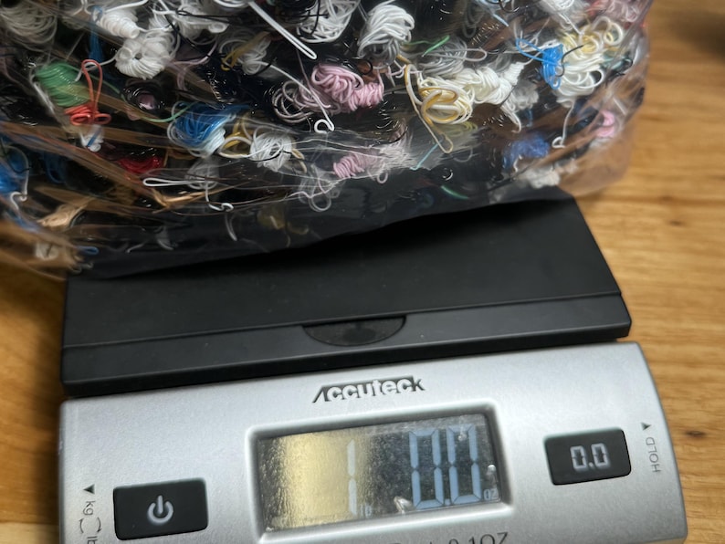 May include: A digital scale with a black base and a silver top. The scale has a digital display that reads "0.0". A clear plastic bag filled with colorful yarn is on top of the scale.