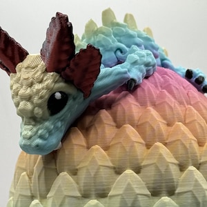 May include: A small, blue and white dragon figurine with red horns, perched on a rainbow-coloured, spiky egg.