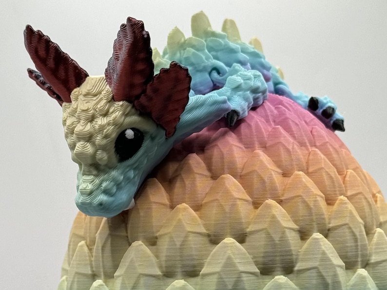 Pu&ograve; includere: Una piccola statuetta di drago blu e bianco con corna rosse, appollaiata su un uovo spinoso arcobaleno.