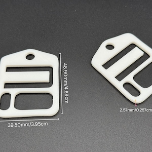 May include: Two white plastic strap adjusters with a rectangular frame, a central horizontal bar, and a circular hole at the top. The dimensions are labeled as 48.90mm by 39.50mm with a thickness of 2.57mm, providing clear technical specifications.