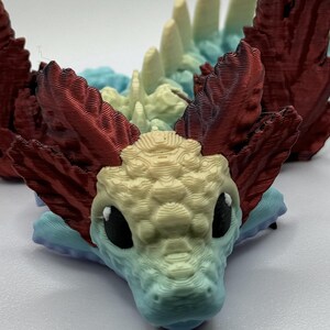 Pu&ograve; includere: Una piccola statuina di drago stampata in 3D, colorata, con squame rosse, blu e gialle. Il drago ha grandi occhi neri e un design testurizzato e dettagliato.