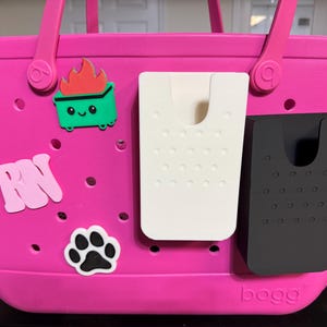 Puede incluir: Un bolso tote rosa brillante con un asa y varios adornos decorativos. Los adornos incluyen un cubo de basura verde con llamas, las letras "RN" y una huella de pata. Dos organizadores de bolsillo están adheridos al lateral del bolso, uno blanco y otro negro.
