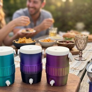 May include: Three colorful beverage coolers with white lids and spouts are displayed on a wooden table. The coolers are teal, purple, and a gradient of purple to green. A can of soda is in a matching purple cooler. The scene suggests an outdoor gathering.