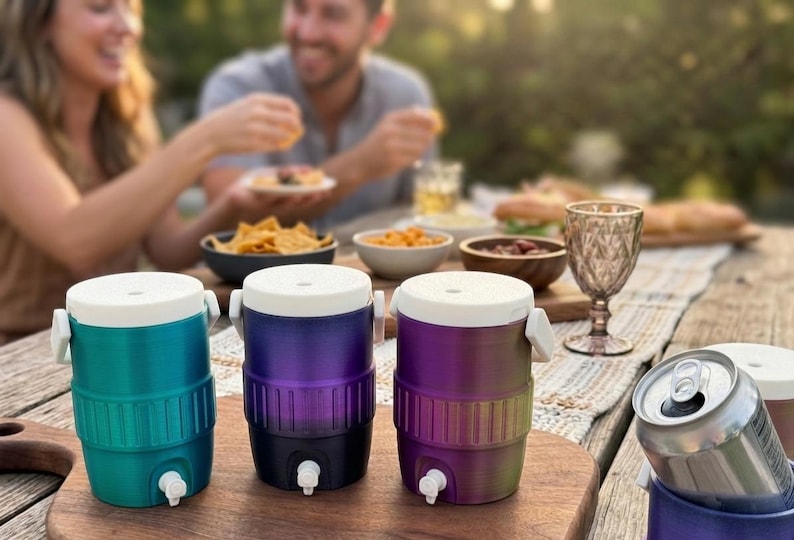 May include: Three colorful beverage coolers with white lids and spouts are displayed on a wooden table. The coolers are teal, purple, and a gradient of purple to green. A can of soda is in a matching purple cooler. The scene suggests an outdoor gathering.