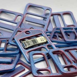 May include: A collection of rectangular plastic buckles with a dual-tone blue and red finish. Each buckle features multiple slots and a circular hole. One buckle in the center displays a small white label with a barcode and the number 211.