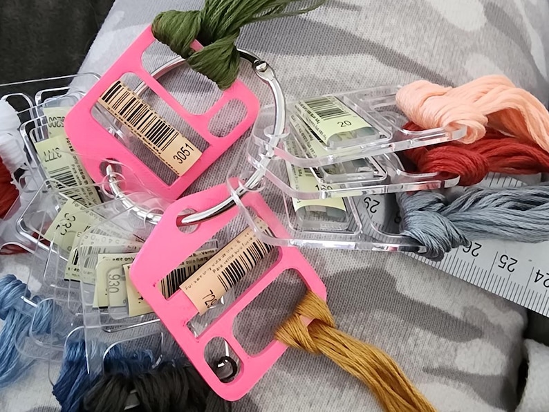 May include: A collection of embroidery floss skeins in various colors attached to clear plastic bobbins and two bright pink plastic organizers on a metal ring. A white measuring tape is visible, showing markings in centimeters and inches.