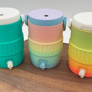 May include: Three colorful water coolers in teal, rainbow, and yellow-orange gradients. Each cooler has a white top, a spigot, and a ribbed band around the middle. The coolers are cylindrical and sit on a wooden surface.
