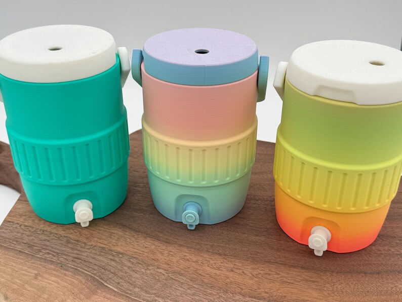 May include: Three colorful water coolers in teal, rainbow, and yellow-orange gradients. Each cooler has a white top, a spigot, and a ribbed band around the middle. The coolers are cylindrical and sit on a wooden surface.