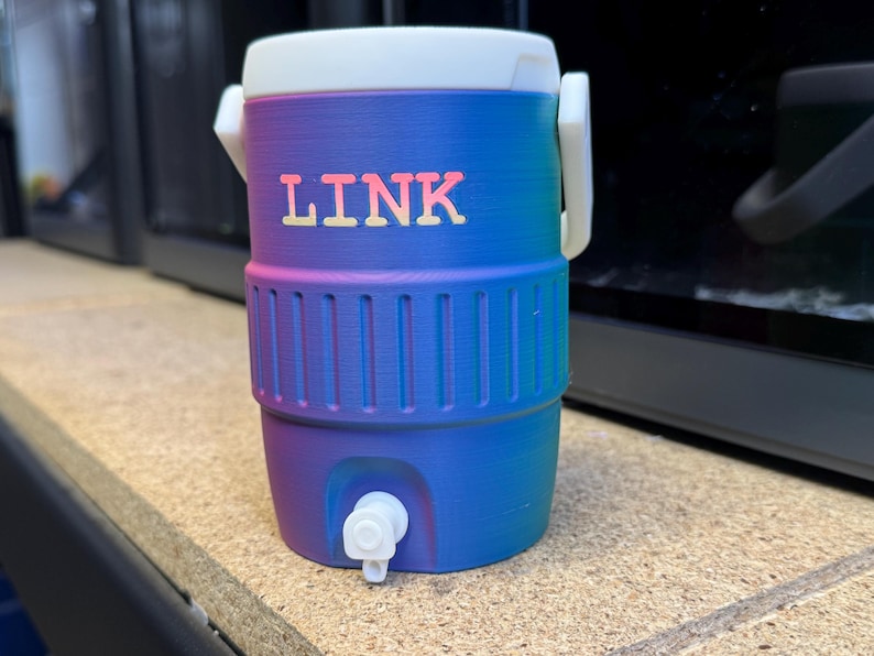 May include: A small, cylindrical cooler with a white lid and handle. The cooler is a gradient of blue and purple with the word "LINK" in orange letters. It has a white spigot at the bottom.