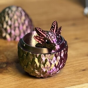 Pu&ograve; includere: Una piccola statuina di drago iridescente &egrave; annidata all'interno di un contenitore a forma di uovo di drago. L'uovo &egrave; realizzato in un materiale metallico con una texture squamosa e uno schema di colori viola e oro.