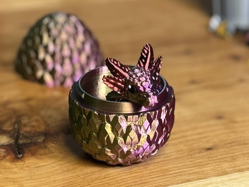 Pu&ograve; includere: Una piccola statuina di drago iridescente &egrave; annidata all'interno di un contenitore a forma di uovo di drago. L'uovo &egrave; realizzato in un materiale metallico con una texture squamosa e uno schema di colori viola e oro.