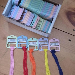 May include: A cardboard box filled with numerous pastel-colored plastic embroidery floss bobbins. Five individual bobbins are arranged in a row, each holding a different colored thread: light pink, red, orange, yellow, and dark blue.