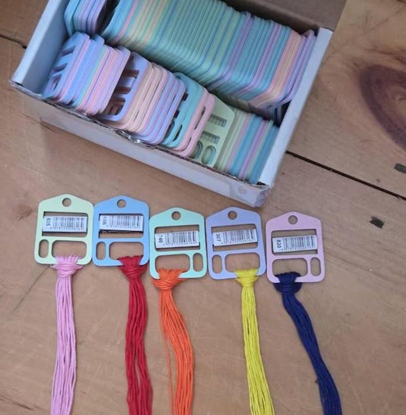 May include: A cardboard box filled with numerous pastel-colored plastic embroidery floss bobbins. Five individual bobbins are arranged in a row, each holding a different colored thread: light pink, red, orange, yellow, and dark blue.