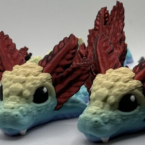 Pu&ograve; includere: Tre figurine stampate in 3D di una creatura mitologica colorata con squame rosse, gialle, blu e verdi. La creatura ha un muso lungo, occhi grandi e una cresta a forma di pinna sulla testa.