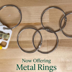 May include: Four silver-colored metal binder rings arranged on a surface. One ring holds a white plastic organizer tab with the number 973 and a barcode printed on it, along with yellow thread. The text Now Offering Metal Rings is visible.