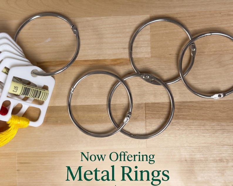 May include: Four silver-colored metal binder rings arranged on a surface. One ring holds a white plastic organizer tab with the number 973 and a barcode printed on it, along with yellow thread. The text Now Offering Metal Rings is visible.