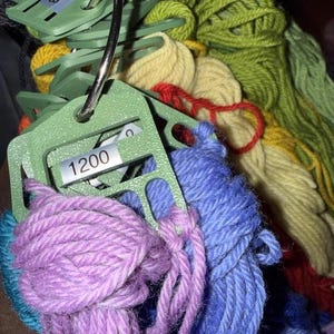 May include: A collection of multi-colored yarn skeins organized on light green plastic bobbins held together by a metal ring. Each bobbin features a small white label with black text, including the number 1200, to identify the specific yarn color.