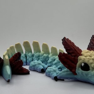 Pu&ograve; includere: Una statuetta di drago stampata in 3D con un corpo segmentato, che presenta un gradiente di colori dal blu chiaro al giallo al rosso. Il drago ha una piccola testa dettagliata con la bocca chiusa e grandi occhi neri.