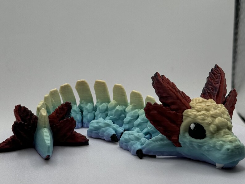 Pu&ograve; includere: Una statuetta di drago stampata in 3D con un corpo segmentato, che presenta un gradiente di colori dal blu chiaro al giallo al rosso. Il drago ha una piccola testa dettagliata con la bocca chiusa e grandi occhi neri.