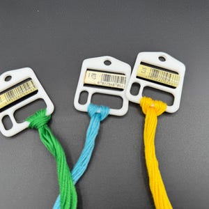 May include: Three white plastic embroidery floss bobbins, each holding a different color of thread: green, light blue, and yellow. Each bobbin features a small rectangular label with a barcode and the numbers 700, 807, and 973 respectively.