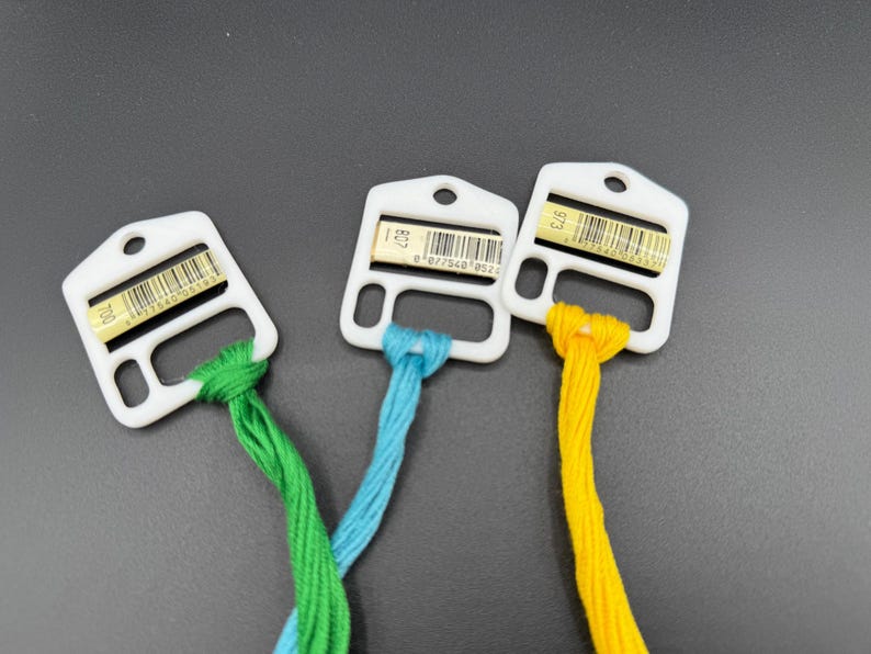 May include: Three white plastic embroidery floss bobbins, each holding a different color of thread: green, light blue, and yellow. Each bobbin features a small rectangular label with a barcode and the numbers 700, 807, and 973 respectively.