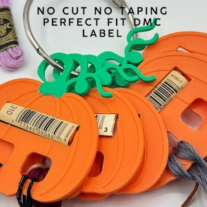May include: A set of orange pumpkin-shaped plastic embroidery floss bobbins on a metal ring, featuring green stems and slots for DMC labels. The text NO CUT NO TAPING PERFECT FIT DMC LABEL is printed above the bobbins, with a skein of purple thread.