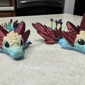 Pu&ograve; includere: Due piccole figurine di drago blu e rosso stampate in 3D con accenti bianchi. I draghi hanno grandi occhi espressivi e sono in posa con le ali spiegate.