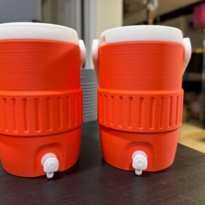 May include: Two orange and white water coolers with spouts. The coolers have a cylindrical shape with a white lid and handle. The lower section of the cooler has vertical ridges. The spouts are white. The coolers are on a dark surface.