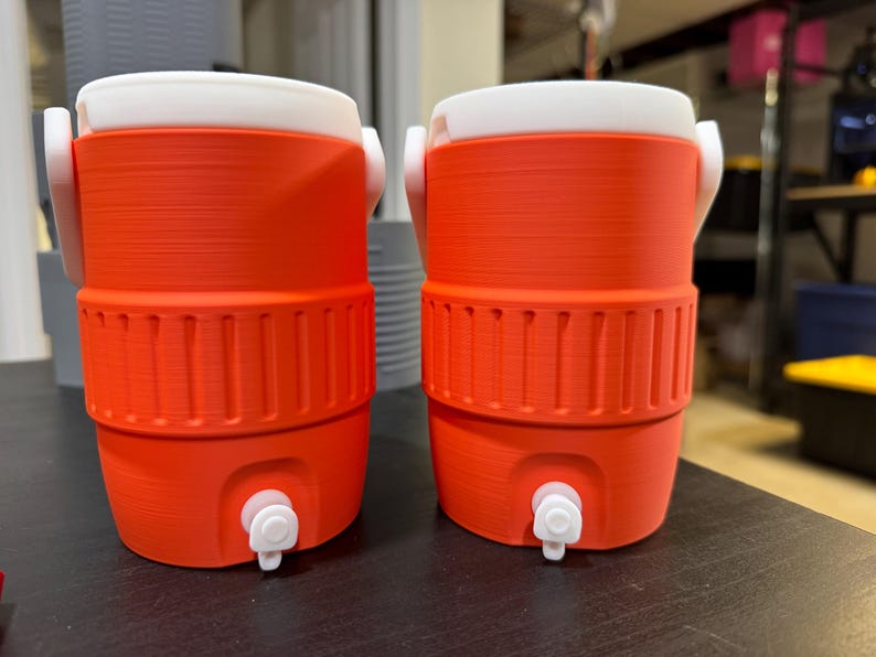 May include: Two orange and white water coolers with spouts. The coolers have a cylindrical shape with a white lid and handle. The lower section of the cooler has vertical ridges. The spouts are white. The coolers are on a dark surface.
