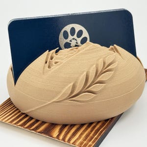 May include: A beige, 3D-printed card holder with a mountain and leaf design. A dark blue card with a paw print logo is held in the holder. The holder sits on a small, wooden base with a striped pattern.