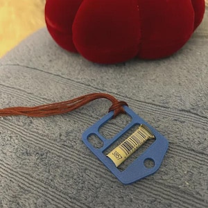 May include: A blue plastic thread organizer with a rectangular cutout and a circular hole, featuring a white paper label with a barcode and the number 3857. A brown embroidery thread is looped through the top of the organizer.