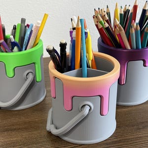 Puede incluir: Tres portalápices grises con diseños de goteo de pintura colorida. Uno tiene un goteo verde, otro un degradado rosa y naranja, y el tercero un goteo morado. Cada recipiente contiene bolígrafos, lápices y rotuladores.