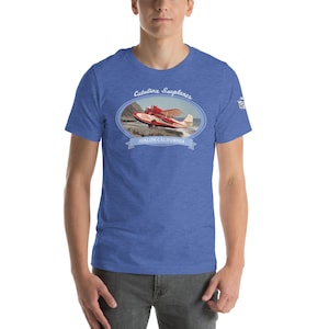 Puede incluir: Una camiseta azul con un gráfico de un hidroavión rojo y blanco despegando del agua. El gráfico está rodeado de un óvalo azul con el texto "Catalina Seaplanes" y "Avalon California".